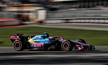 Colorful Formula 1 car with BWT livery speeding along a race track, motion blur background behind it.