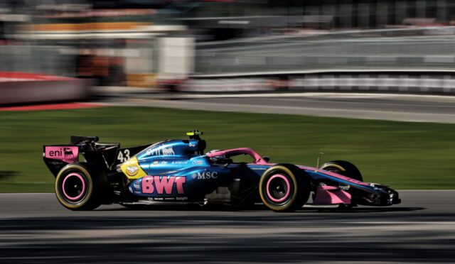 Colorful Formula 1 car with BWT livery speeding along a race track, motion blur background behind it.