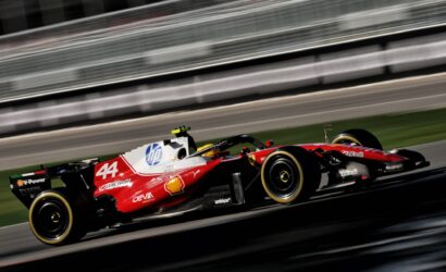 Red Formula 1 race car speeding along a track with sponsor logos visible and motion blur in the background.