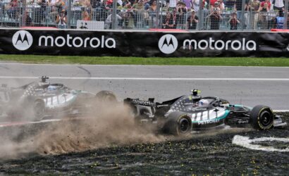 Two Formula 1 cars crash on a gravel runoff, dust and debris spraying as spectators watch behind a chain-link fence and a Motorola barrier.”,”F1 cars collide near the track edge, kicking up dirt while fans photograph from behind the fence and sponsor banners.”,”Race cars in a high-speed crash on the runoff area, crowd watching intently from behind the fence.