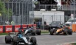 Formula 1 race cars speeding along a fenced track with red/white barriers and Lenovo banners, spectators watching from the stands above the pit area.