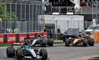 Formula 1 race cars speeding along a fenced track with red/white barriers and Lenovo banners, spectators watching from the stands above the pit area.