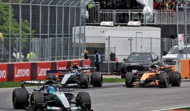Formula 1 race cars speeding along a fenced track with red/white barriers and Lenovo banners, spectators watching from the stands above the pit area.