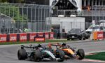 Two Formula 1 cars race side by side on a track with red Lenovo barriers and a crowded grandstand in the background.