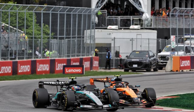 Two Formula 1 cars race side by side on a track with red Lenovo barriers and a crowded grandstand in the background.