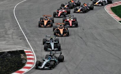 Multiple Formula 1 cars race in a tight group around a curved track, sponsor logos visible on the cars.
