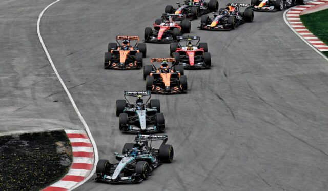 Multiple Formula 1 cars race in a tight group around a curved track, sponsor logos visible on the cars.