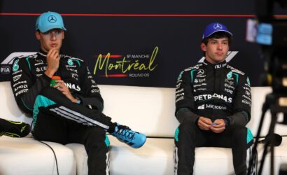 Two Mercedes F1 drivers sit on a white sofa during a press event, wearing team suits and blue caps, with a Montreal backdrop behind them and a red line above the logo.
