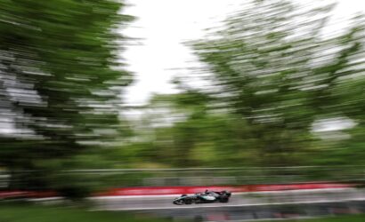 Formula-style race car speeding on a racetrack with red barriers and blurred green trees in the background.