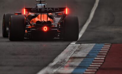 Rear view of an orange Formula 1 car on track, red tail lights glowing, curbs painted blue, white, and red on the right edge.