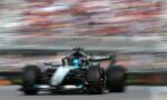 Motion-blurred Formula 1 car speeding toward the camera on a gray racetrack with a colorful, blurred grandstand background.