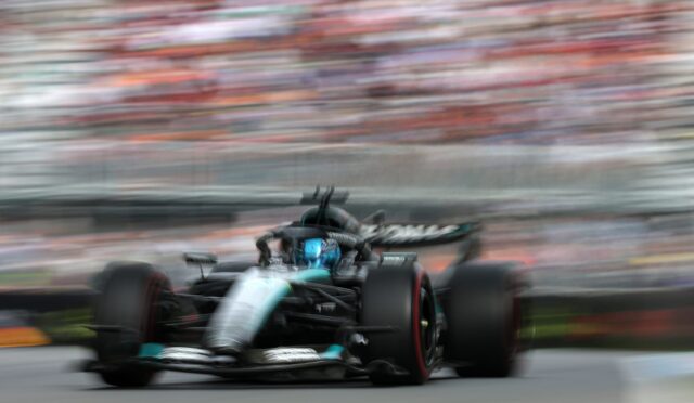Motion-blurred Formula 1 car speeding toward the camera on a gray racetrack with a colorful, blurred grandstand background.