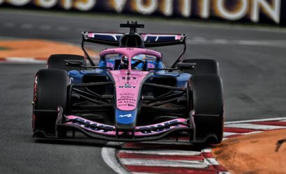 Front-on view of a pink-and-blue Formula 1 car taking a tight turn on a racetrack, wheels in motion.