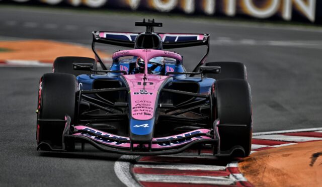 Front-on view of a pink-and-blue Formula 1 car taking a tight turn on a racetrack, wheels in motion.