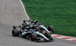 Three Formula 1 race cars hug a left-hand corner on a track, with green grass beside the asphalt (Mercedes‑AMG livery).