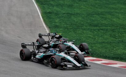Three Formula 1 race cars hug a left-hand corner on a track, with green grass beside the asphalt (Mercedes‑AMG livery).