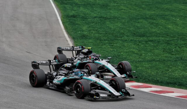 Three Formula 1 race cars hug a left-hand corner on a track, with green grass beside the asphalt (Mercedes‑AMG livery).