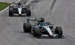Two Mercedes Formula 1 cars race on a gray track, the front car leading with teal accents and the second car close behind.