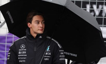 Young man in a dark racing jacket with sponsor logos stands under a large black umbrella at a race paddock or pit area.