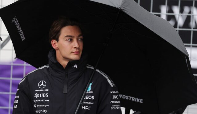 Young man in a dark racing jacket with sponsor logos stands under a large black umbrella at a race paddock or pit area.