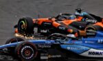 Close-up of several Formula 1 cars tightly packed on the track, showing orange and blue liveries and sponsor logos with sparks near the tires.