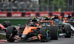 Orange Formula 1 car leads a pack on a race track with blurred spectators in the background.
