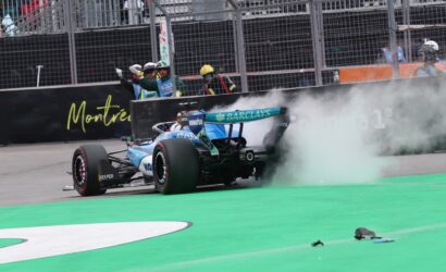 Blue Formula 1 car drifting with smoke near the pit wall, crew signaling in the background on a race track in Montreal.",