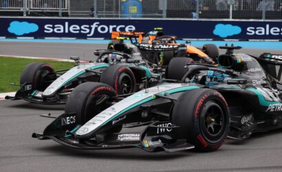 Three Formula 1 cars in a tight race, Mercedes–Petronas teal livery leading on a track with blue barriers in the background