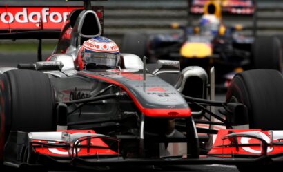 Front view of a silver Formula 1 car on the track, Vodafone branding on the helmet and car, with a Red Bull car blurred in the background