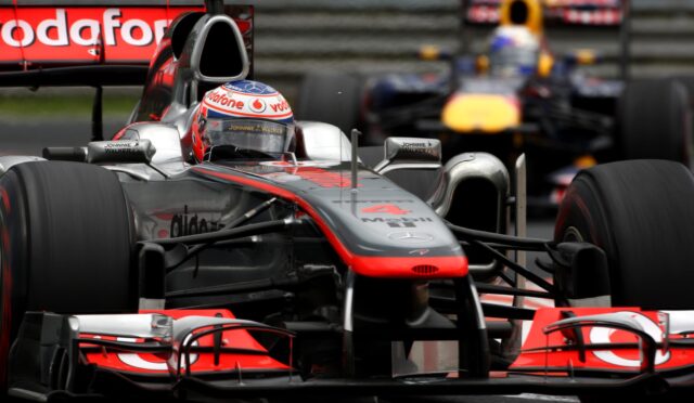 Front view of a silver Formula 1 car on the track, Vodafone branding on the helmet and car, with a Red Bull car blurred in the background