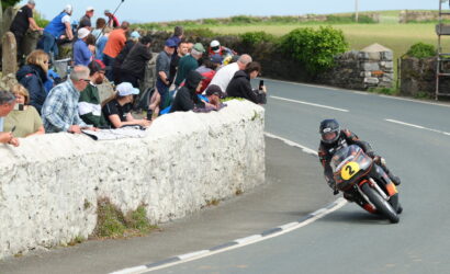 Motorcycle racer leaning into a sharp turn on a rural road as a crowd watches from a stone wall beside the track, bike bearing number 2.