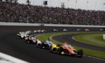 Racing cars on a crowded oval track turning left, with a packed stadium of spectators above.