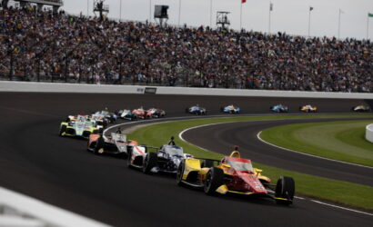 Racing cars on a crowded oval track turning left, with a packed stadium of spectators above.