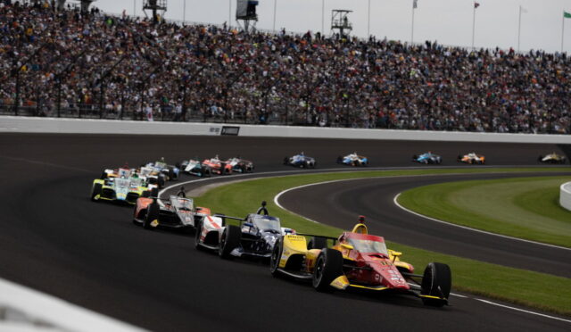 Racing cars on a crowded oval track turning left, with a packed stadium of spectators above.