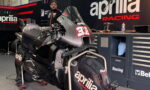 Race bike on a stand inside a garage, branded with Aprilia Racing logos and a large number plate on the front fairing, with a mechanic in the background and tool chests nearby.