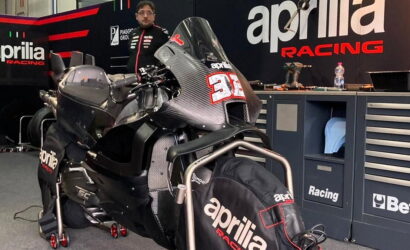 Race bike on a stand inside a garage, branded with Aprilia Racing logos and a large number plate on the front fairing, with a mechanic in the background and tool chests nearby.