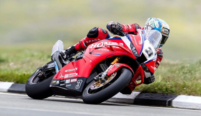 Moto racer in a red Honda race bike carving through a left-hand turn on a track, leaning aggressively with a blue-white helmet nearby.