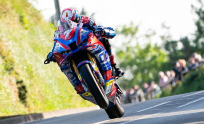 Motorcycle racer in colorful suit and helmet leaning into a turn on a road, with spectators in the background.