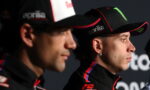 Two racers in black caps and racing jackets, facing right, during a press event with sponsor logos on hats (Aprilia, Monster).