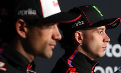 Two racers in black caps and racing jackets, facing right, during a press event with sponsor logos on hats (Aprilia, Monster).