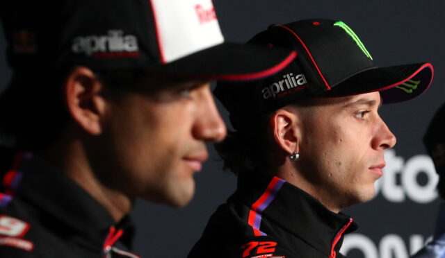 Two racers in black caps and racing jackets, facing right, during a press event with sponsor logos on hats (Aprilia, Monster).