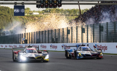 Two race cars sprint toward the camera on a racing circuit as sparks fly from the barrier and a grandstand fills the background