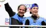 Two smiling men on a podium celebrate a victory; one in a blue-and-black jacket raises a trophy while a young racer in blue gear stands beside him.
