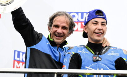 Two smiling men on a podium celebrate a victory; one in a blue-and-black jacket raises a trophy while a young racer in blue gear stands beside him.
