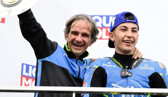 Two smiling men on a podium celebrate a victory; one in a blue-and-black jacket raises a trophy while a young racer in blue gear stands beside him.
