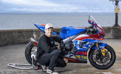 Man kneeling beside a blue, red, and white racing motorcycle on a sea-side pier, posing for the photo.