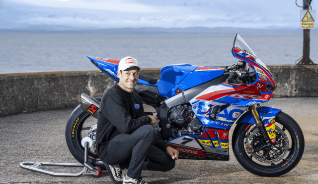 Man kneeling beside a blue, red, and white racing motorcycle on a sea-side pier, posing for the photo.