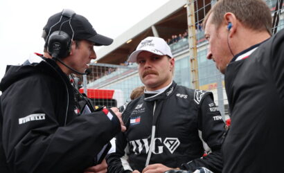 Race car driver in black racing suit and white cap stands between two team members in a pit area, listening intently.