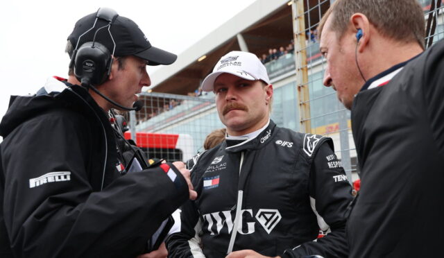 Race car driver in black racing suit and white cap stands between two team members in a pit area, listening intently.