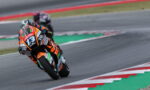 Racer in orange-and-black suit leans into a sharp turn on a race track, bike number 13 adorned with sponsor stickers.